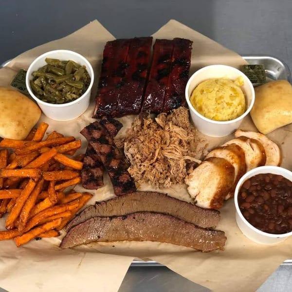 A large platter of Doc's Smokehouse BBQ with ribs, pulled pork, brisket, turkey, sweet potato fries, green beans, corn pudding, baked beans, and rolls.
