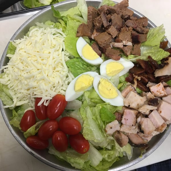 A large salad with lettuce, shredded cheese, cherry tomatoes, hard-boiled eggs, and various smoked meats.